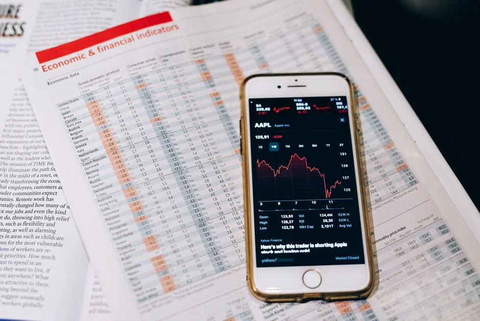 Hands holding a smartphone next to a laptop displaying stock charts, illustrating digital trading.